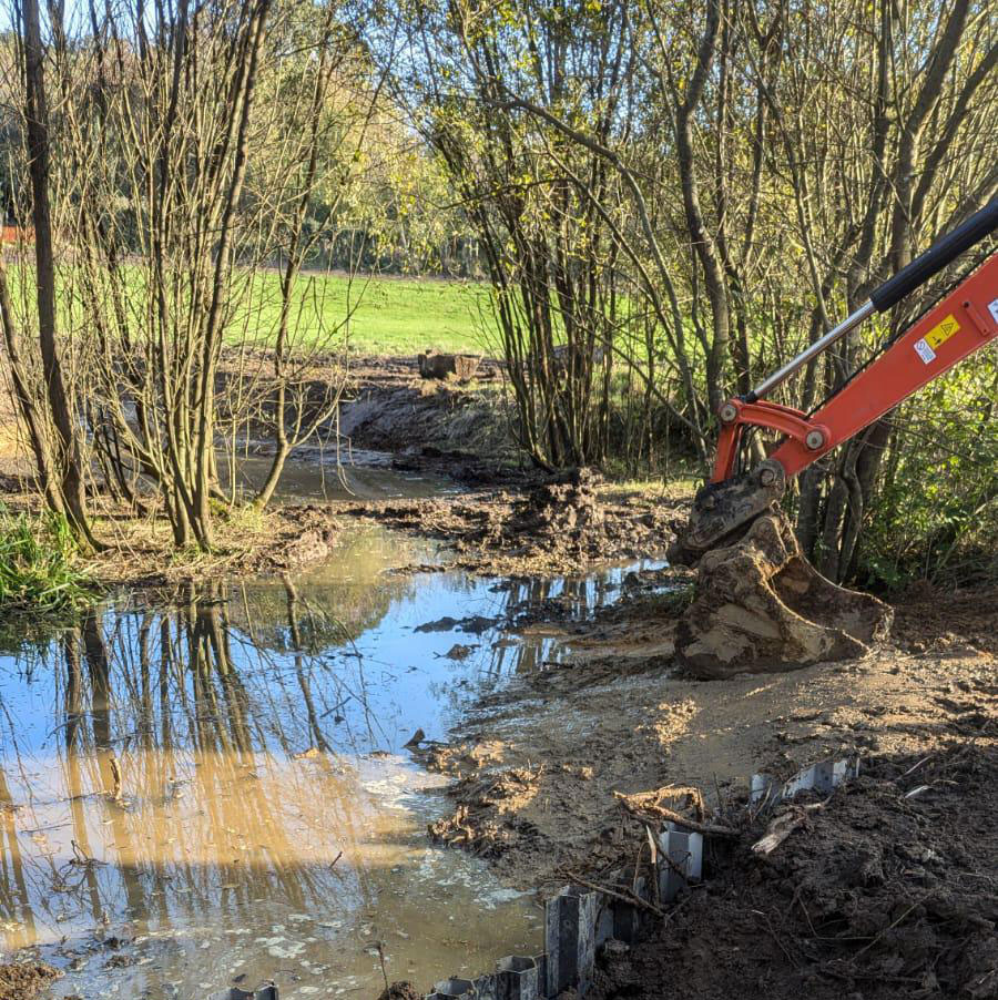 A compact excavator shaping the muddy margin of a new pond, with water reflecting bare trees and open pasture beyond — wetland creation in progress.
